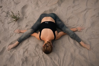 A person is performing a yoga stretch on a sandy surface, with legs outstretched to the sides and arms extended forward. The person is wearing a black top and gray leggings, and the hair is tied back.