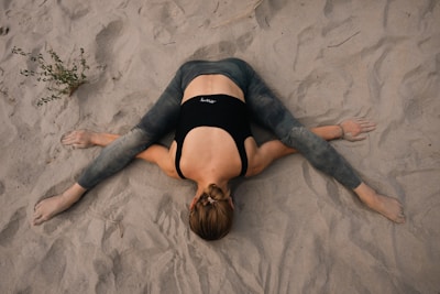 A person is performing a yoga stretch on a sandy surface, with legs outstretched to the sides and arms extended forward. The person is wearing a black top and gray leggings, and the hair is tied back.