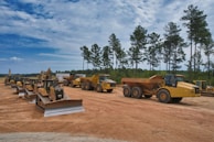 A line of large construction vehicles, including bulldozers and dump trucks, is parked on a dirt clearing. The vehicles are a vivid yellow, contrasting with the earthy brown of the ground. Tall pine trees and a mostly cloudy blue sky form the background, creating a serene and orderly construction scene.