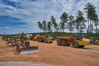 A line of large construction vehicles, including bulldozers and dump trucks, is parked on a dirt clearing. The vehicles are a vivid yellow, contrasting with the earthy brown of the ground. Tall pine trees and a mostly cloudy blue sky form the background, creating a serene and orderly construction scene.