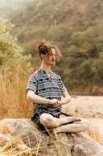 A person with curly hair is sitting in a meditative pose on a rock surrounded by tall grass. The background shows a blurred forested area with muted tones, suggesting a serene and natural environment.