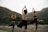 Three people are practicing yoga by a river, standing on rocks in a tree pose with their hands in the air. They are surrounded by a natural landscape with mountains and greenery in the background.