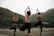 Three people are practicing yoga by a river, standing on rocks in a tree pose with their hands in the air. They are surrounded by a natural landscape with mountains and greenery in the background.