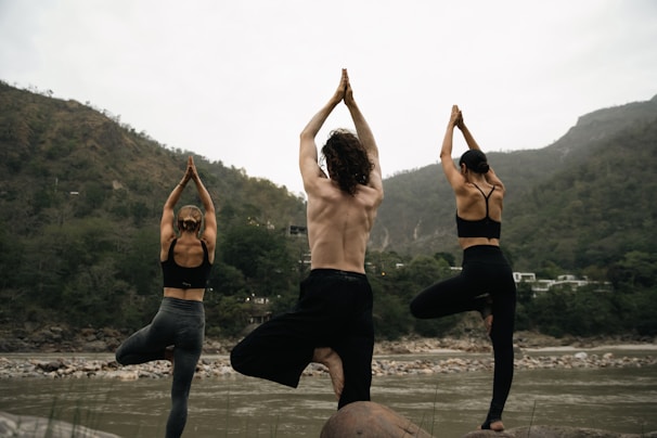 Three people practicing yoga by the Ganges River in Rishikesh with the Himalayas in the background