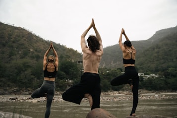 Three people are practicing yoga by a river, standing on rocks in a tree pose with their hands in the air. They are surrounded by a natural landscape with mountains and greenery in the background.