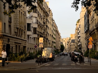 An urban street scene lined with classic, beige-toned buildings displaying French architectural style. Vehicles are parked along the road, with a van prominently in view. Pedestrians and cyclists are present, and traffic signs, including speed and pedestrian zone signs, are visible. Lush greenery partially covers the building facades, and the street is bathed in the soft light of either early morning or late afternoon.