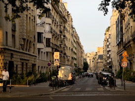 An urban street scene lined with classic, beige-toned buildings displaying French architectural style. Vehicles are parked along the road, with a van prominently in view. Pedestrians and cyclists are present, and traffic signs, including speed and pedestrian zone signs, are visible. Lush greenery partially covers the building facades, and the street is bathed in the soft light of either early morning or late afternoon.