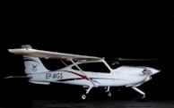 A small white airplane with a single propeller is situated in a dark hangar. The aircraft has an identification code 'EP-MGS' on its side and features sleek blue and red stripes as part of its design.