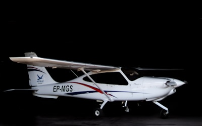A small white airplane with a single propeller is situated in a dark hangar. The aircraft has an identification code 'EP-MGS' on its side and features sleek blue and red stripes as part of its design.
