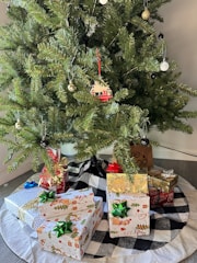 A decorated Christmas tree with various ornaments, including round baubles and a small house hanging. Below the tree, there are several wrapped gifts with colorful bows, placed on a black and white checkered tree skirt.