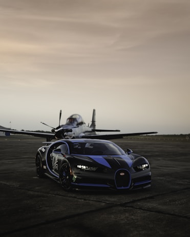 A sleek, black and blue sports car is parked on a runway in front of a small military aircraft. The setting sun casts a soft, warm light over the scene, highlighting the smooth curves of the car and the aerodynamic design of the plane. The ground is dark and slightly damp, indicating a recent rain.