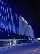 A modern building with the name 'GHELAMCO ARENA' illuminated in bright lights. The façade consists of vertical blue light strips, creating a vibrant and sleek appearance. The lower level has glass windows displaying the logo 'energylab', and a few people are visible on the sidewalk.