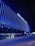 A modern building with the name 'GHELAMCO ARENA' illuminated in bright lights. The façade consists of vertical blue light strips, creating a vibrant and sleek appearance. The lower level has glass windows displaying the logo 'energylab', and a few people are visible on the sidewalk.