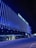 A modern building with the name 'GHELAMCO ARENA' illuminated in bright lights. The façade consists of vertical blue light strips, creating a vibrant and sleek appearance. The lower level has glass windows displaying the logo 'energylab', and a few people are visible on the sidewalk.
