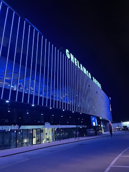 A modern building with the name 'GHELAMCO ARENA' illuminated in bright lights. The fa&ccedil;ade consists of vertical blue light strips, creating a vibrant and sleek appearance. The lower level has glass windows displaying the logo 'energylab', and a few people are visible on the sidewalk.