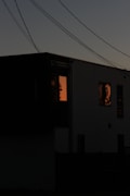 A dark silhouette of a modern house against a twilight sky. There are visible power lines cutting across the top of the image. The glow of a warm, orange light is seen through two windows, suggesting interior illumination.