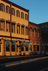 A historic building with ornate architectural details, including large windows, decorative moldings, and warm tones, bathed in the golden light of the setting sun. A hanging flower basket is attached to a vintage-style street lamp in front.