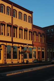 A historic building with ornate architectural details, including large windows, decorative moldings, and warm tones, bathed in the golden light of the setting sun. A hanging flower basket is attached to a vintage-style street lamp in front.