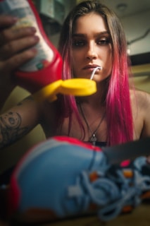 A person with pink-dyed hair is drinking ketchup through a straw. The scene also features colorful sneakers in the foreground. Tattoos are visible on the person's arm, and they are wearing a necklace.