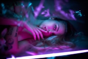 A person lies in a relaxed pose under soft, colorful lighting. Their expression is calm and pensive, with one hand resting gently near the face. The lighting creates a dreamy atmosphere with shades of pink and blue.
