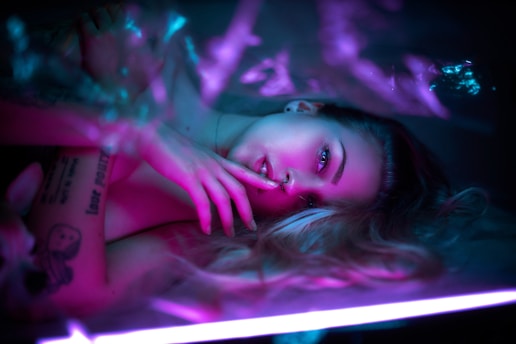 A person lies in a relaxed pose under soft, colorful lighting. Their expression is calm and pensive, with one hand resting gently near the face. The lighting creates a dreamy atmosphere with shades of pink and blue.