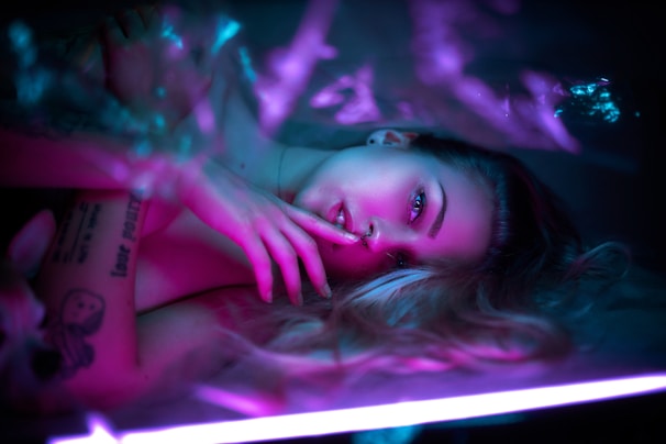 A person lies in a relaxed pose under soft, colorful lighting. Their expression is calm and pensive, with one hand resting gently near the face. The lighting creates a dreamy atmosphere with shades of pink and blue.