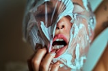A person with a plastic sheet wrapped around their face, partially covering the mouth and nose. The mouth is open and part of the lips and teeth are visible. The hand is gripping the plastic with painted nails.