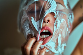 A person with a plastic sheet wrapped around their face, partially covering the mouth and nose. The mouth is open and part of the lips and teeth are visible. The hand is gripping the plastic with painted nails.