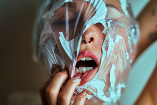 A person with a plastic sheet wrapped around their face, partially covering the mouth and nose. The mouth is open and part of the lips and teeth are visible. The hand is gripping the plastic with painted nails.