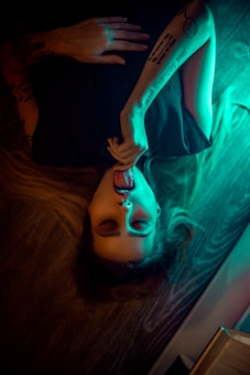 A person lies on their back with closed eyes and a relaxed expression. The lighting casts a blue and green glow, highlighting tattoos on the arm. The scene is dimly lit, creating an artistic and moody atmosphere.