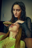 A person with long dark hair pours a liquid into the mouth of another person with light brown hair, who is wearing a neon green mesh top. The scene appears to be intimate, with the second person lying back and having their eyes closed. The background is dimly lit, giving a focused emphasis on the two individuals.