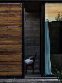 A modern architectural scene featuring a partly open sliding door with wooden slats on the left and a large glass window on the right. Beyond the window is a glimpse of an interior space with a chair partially covered by a pillow and fabric. The background reveals concrete walls and a hint of vegetation outside. The sky outside is tinged with warm sunset hues.
