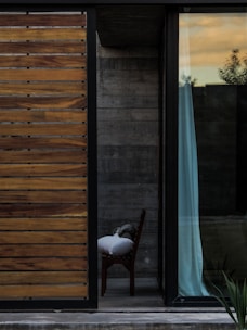 A modern architectural scene featuring a partly open sliding door with wooden slats on the left and a large glass window on the right. Beyond the window is a glimpse of an interior space with a chair partially covered by a pillow and fabric. The background reveals concrete walls and a hint of vegetation outside. The sky outside is tinged with warm sunset hues.