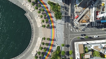 An aerial view of an urban waterfront area features a wide, curved promenade next to a body of water. A colorful rainbow pathway adds vibrancy along the sidewalk, accompanied by evenly spaced green trees. Adjacent to this area is a busy intersection with cars, sidewalks, and construction sites, bordered by various buildings.