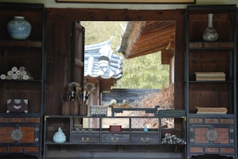 The image depicts a traditional interior space with wooden shelves and drawers, housing various items such as rolled-up scrolls, brushes in a holder, ceramic vases, and small decorative boxes. Through a window or opening, an ornate roof with blue tiles is visible, hinting at a traditional architectural style surrounded by greenery.