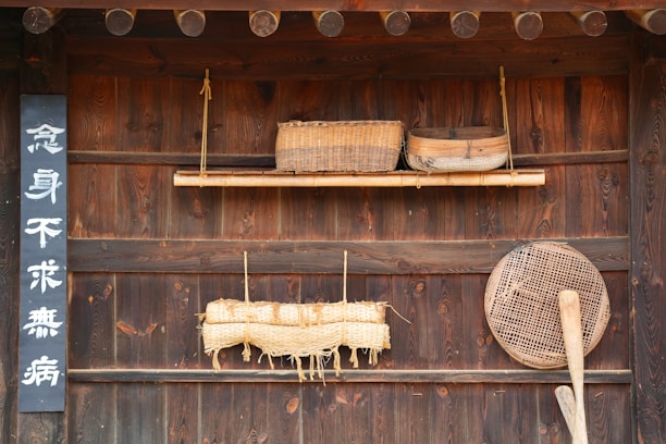 A wooden wall decorated with traditional items, including a woven basket, a wooden tray, a straw mat tied with rope, and a large wooden paddle next to a woven sieve. There is also a vertical sign with white characters on a black background.