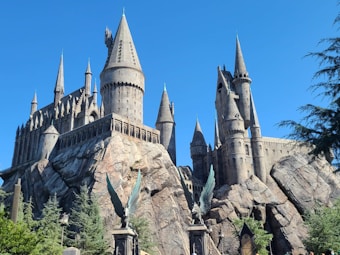 A majestic castle with multiple spires and towers sits atop rocky cliffs surrounded by lush greenery. The architecture is gothic-themed with intricate stone detailing. Statues of winged creatures are prominently displayed at the forefront, adding a mystical element to the scene. The sky is clear and blue, enhancing the grandeur of the structure.