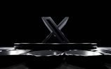 A large, metallic 'X' stands prominently on a glossy, black, platform surrounded by reflective, polygonal surfaces. The setting is dark with dramatic lighting highlighting the contours of the 'X'.