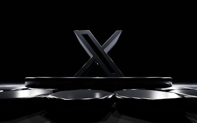 A large, metallic 'X' stands prominently on a glossy, black, platform surrounded by reflective, polygonal surfaces. The setting is dark with dramatic lighting highlighting the contours of the 'X'.