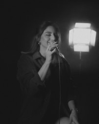 A person is singing into a microphone, illuminated by a bright light source behind them. The setting appears to be intimate, conveying a sense of performance or rehearsal. The image is in black and white, focusing on the subject who seems to be enjoying the moment.