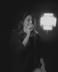 A person is singing into a microphone, illuminated by a bright light source behind them. The setting appears to be intimate, conveying a sense of performance or rehearsal. The image is in black and white, focusing on the subject who seems to be enjoying the moment.