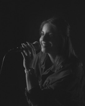 A black and white photograph of a woman holding a microphone, appearing to be singing or speaking. The scene is dimly lit, creating a moody atmosphere with soft shadows and highlights across her face. She is wearing a watch on her left wrist and has long hair, framing her expression.