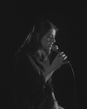 A black and white photograph of a person singing into a microphone. The individual has long hair and is wearing a dark jacket. The background is entirely black, creating a focused spotlight effect on the singer.