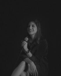 A person sits with a relaxed pose holding a microphone, smiling softly. The setting is monochrome with a dark background, creating a calm atmosphere. They are wearing a watch and have long hair.