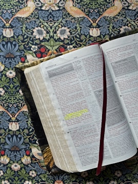 An open Bible lies on a floral patterned surface, with passages highlighted in yellow. The pages show sections with red text, indicating words of significance, and a red ribbon bookmark is placed between the pages. The background features an ornate design with birds, flowers, and leaves.