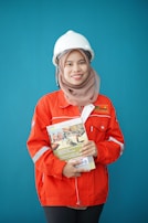 A person wearing a bright red industrial jacket with reflective stripes and a white safety helmet. They are holding a book titled 'Occupational Safety and Health Act' against a plain blue background. The person is smiling and the jacket has a logo on the chest.