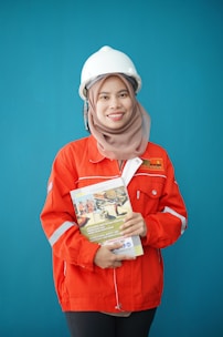 A person wearing a bright red industrial jacket with reflective stripes and a white safety helmet. They are holding a book titled 'Occupational Safety and Health Act' against a plain blue background. The person is smiling and the jacket has a logo on the chest.
