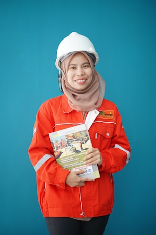 A person wearing a bright red industrial jacket with reflective stripes and a white safety helmet. They are holding a book titled 'Occupational Safety and Health Act' against a plain blue background. The person is smiling and the jacket has a logo on the chest.
