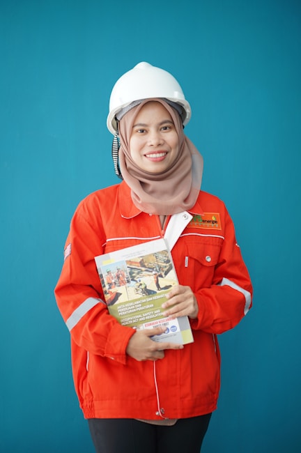 A person wearing a bright red industrial jacket with reflective stripes and a white safety helmet. They are holding a book titled 'Occupational Safety and Health Act' against a plain blue background. The person is smiling and the jacket has a logo on the chest.