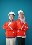 Two individuals are standing back-to-back with arms crossed, wearing red safety jackets and white hard hats. Both are smiling and appear confident. The background is a solid teal color, adding contrast to their bright attire.